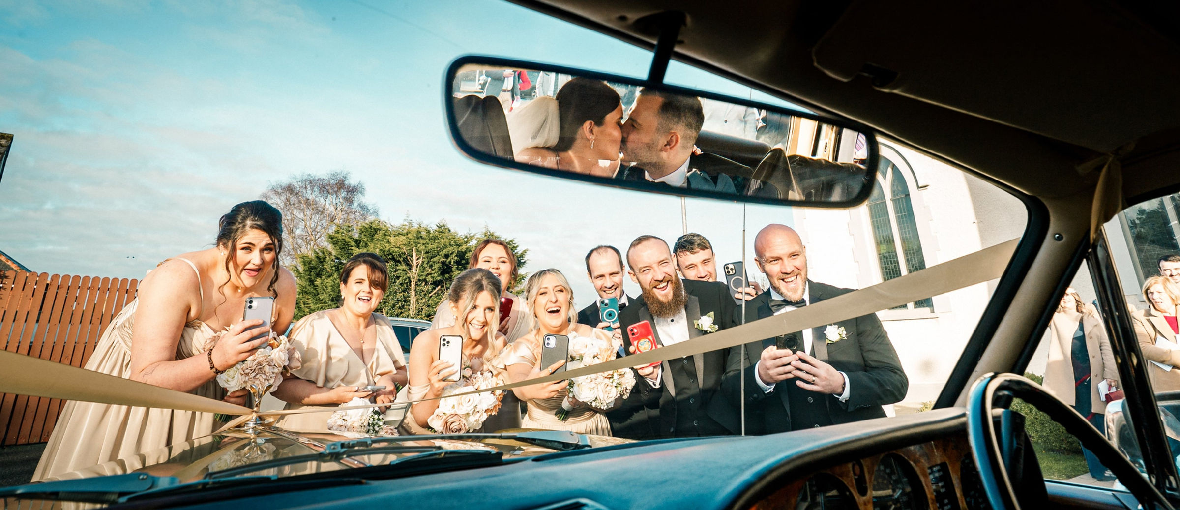 A wedding party laughing around a vintage car while the couple kiss in its mirror